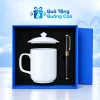 Minh họa cốc sứ trắng quai vuông trong hộp xanh khay đen, nắp cốc nằm riêng phía trên và bút đen bên phải
