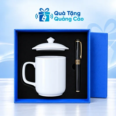 Minh họa cốc sứ trắng quai vuông trong hộp xanh khay đen, nắp cốc nằm riêng phía trên và bút đen bên phải