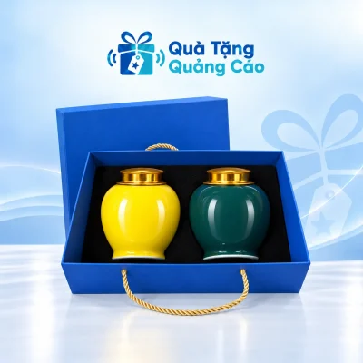 Minh họa hũ chè vàng bên trái và xanh đậm bên phải, nắp vàng kim, cùng hộp xanh khay đen có dây xách
