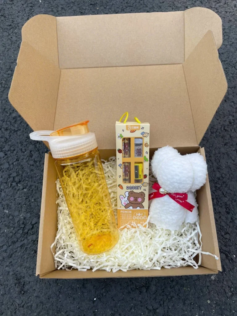 Ảnh tư liệu hộp carton mở với bình nhựa vàng, bộ bút chì tẩy gọt và khăn gấu trắng