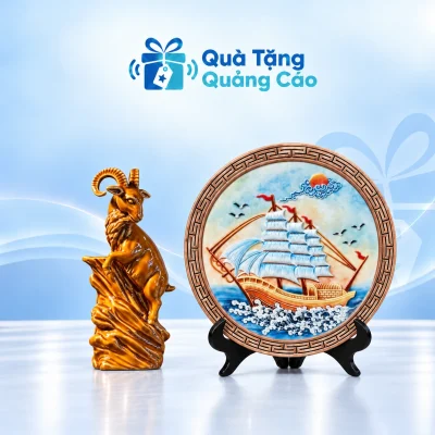 Minh họa tượng dê màu hổ phách và đĩa thuyền buồm kèm kệ gỗ trên nền xanh nhạt Quà Tặng Quảng Cáo