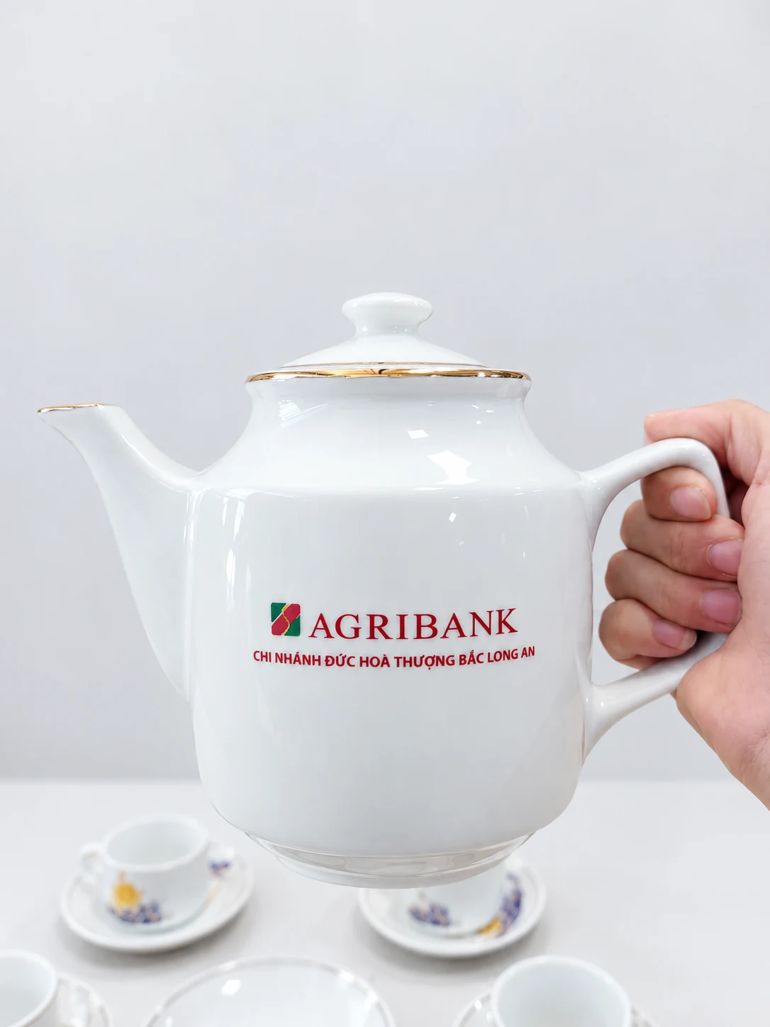 Ấm trắng có nắp, vòi và quai cầm, giữ phần in Agribank ở ảnh nguồn
