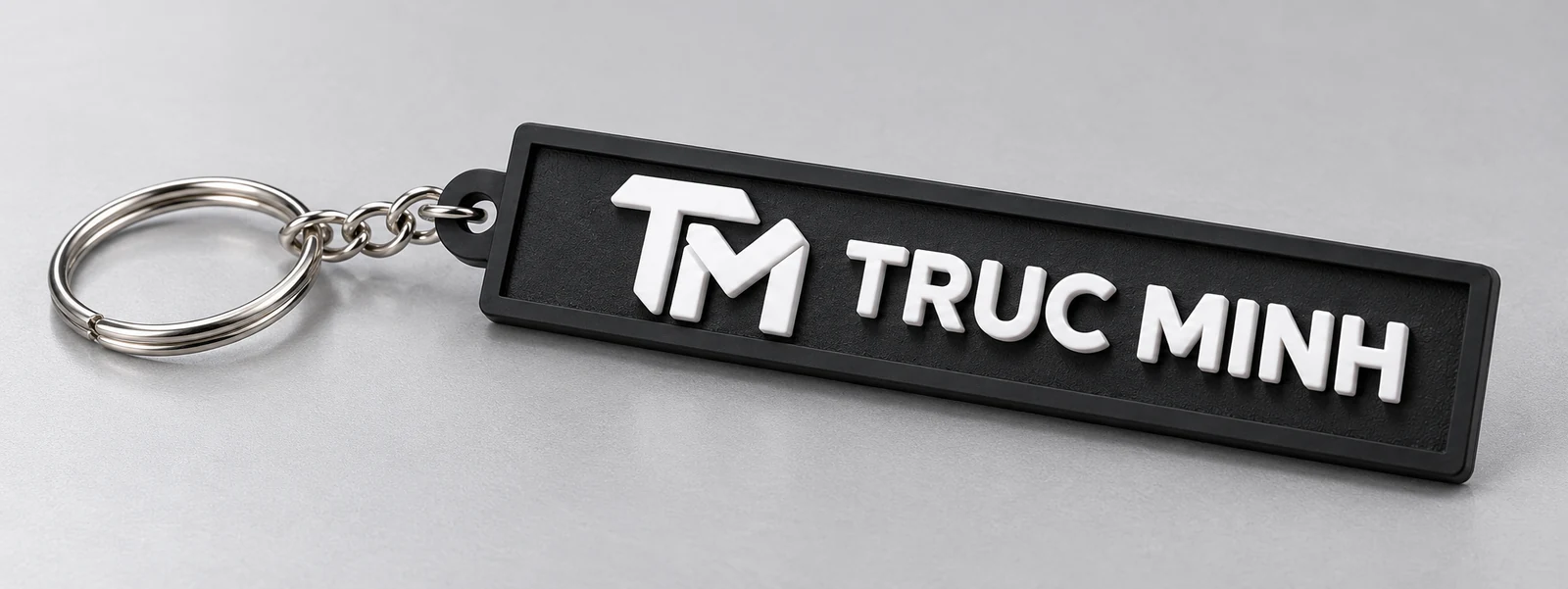 Minh họa chữ nổi TRUC MINH Cận cảnh biểu tượng TM và chữ TRUC MINH nổi trắng trên nền cao su đen có viền chữ nhật