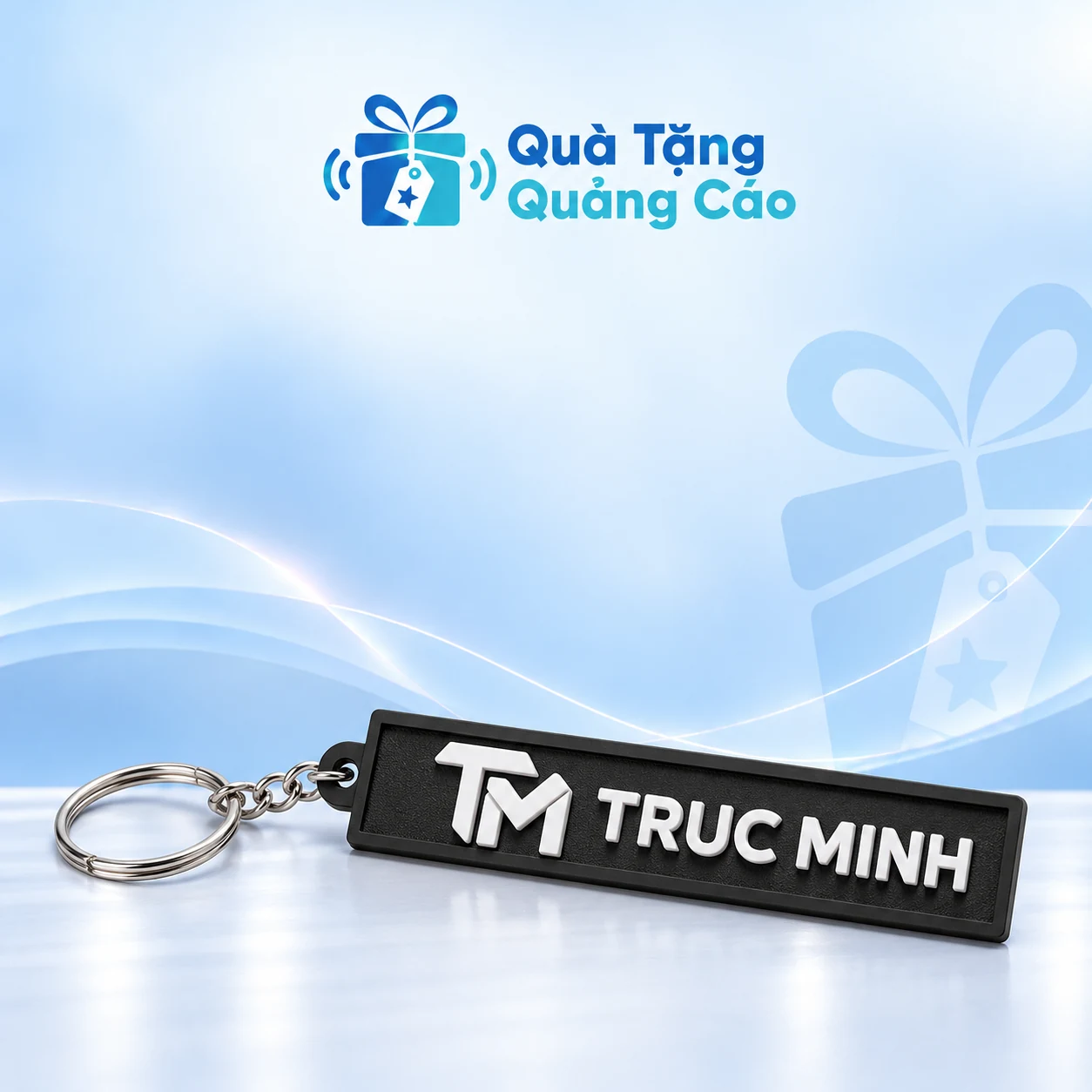 Móc khóa cao su bản dài trên nền xanh nhạt Móc khóa cao su đen bản dài có dây xích, khoen tròn và chữ nổi TRUC MINH trên nền Quà Tặng Quảng Cáo