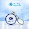 Một móc khóa mica hình tròn viền xanh có khoen tròn, dây xích và nhận diện NHAT HUY trên nền Quà Tặng Quảng Cáo