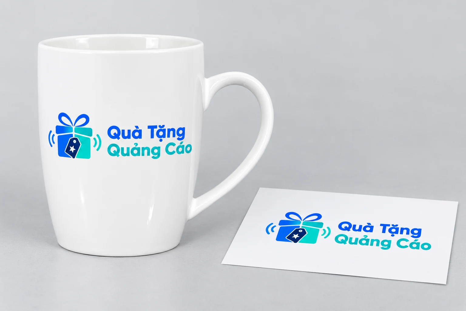 Logo Quà Tặng Quảng Cáo minh họa trên cốc trắng cạnh bản giấy